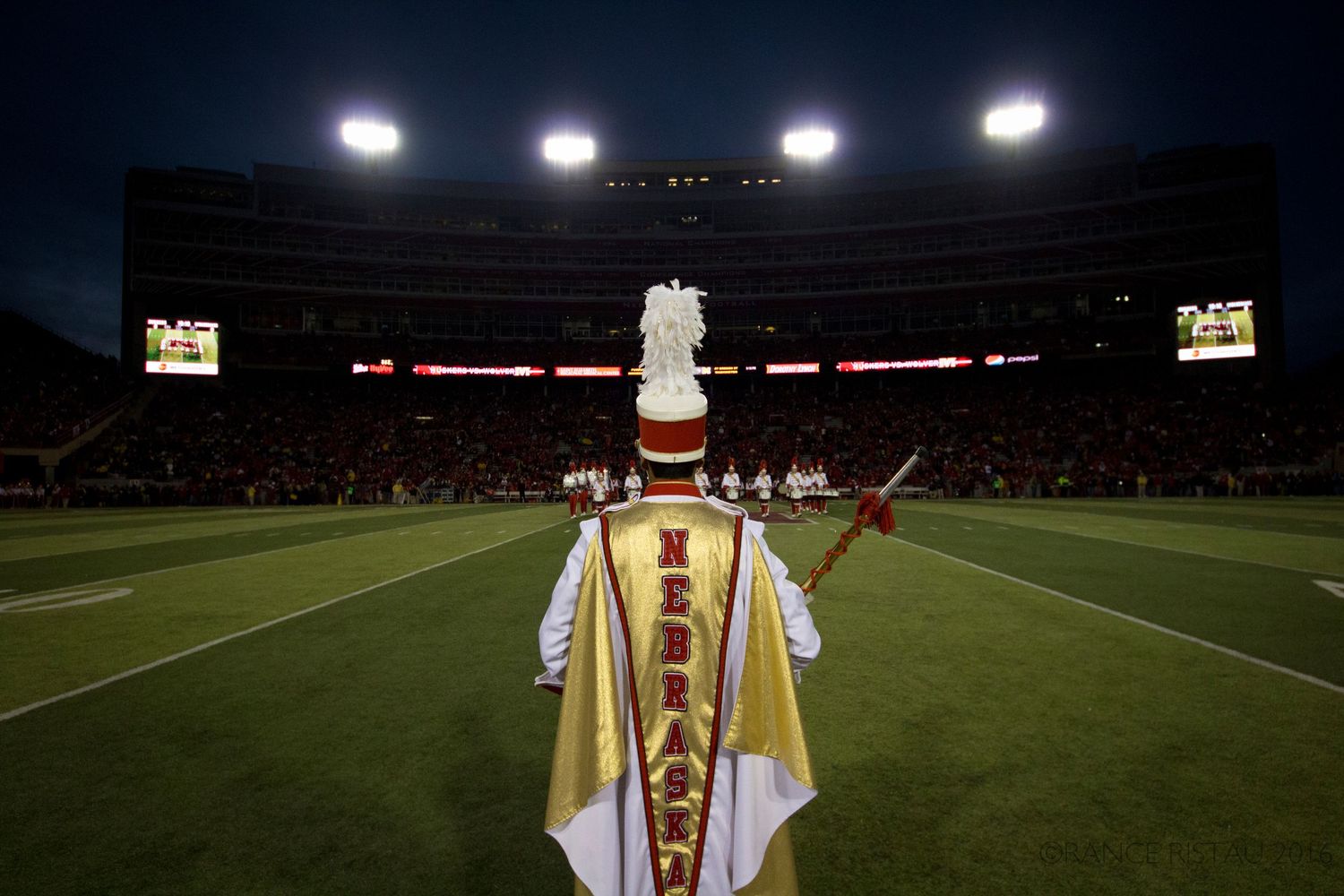 Marching band member in uniform stands on football field under stadium lights.