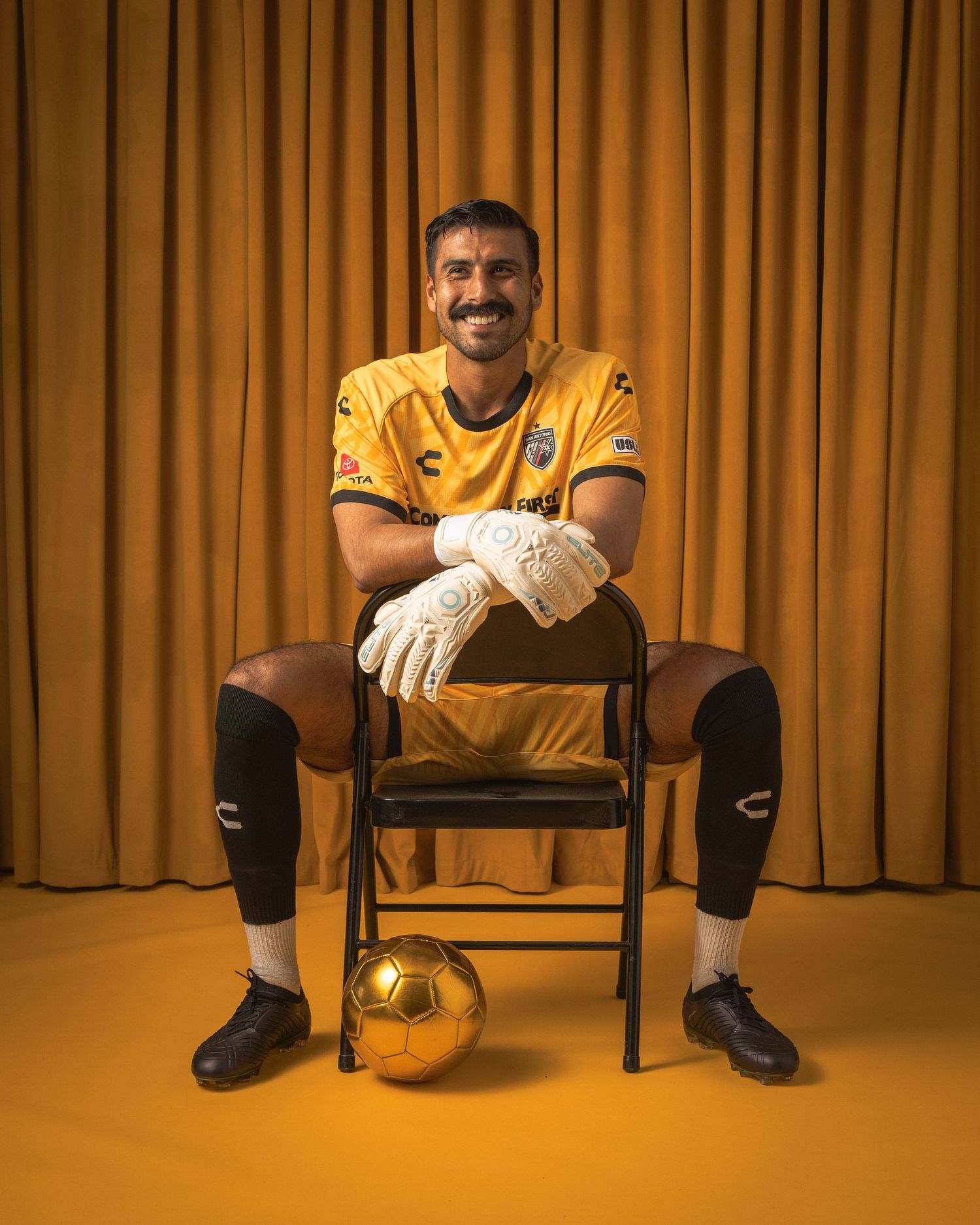 Soccer player in yellow goalkeeper uniform sits on chair with ball against gold curtain.
