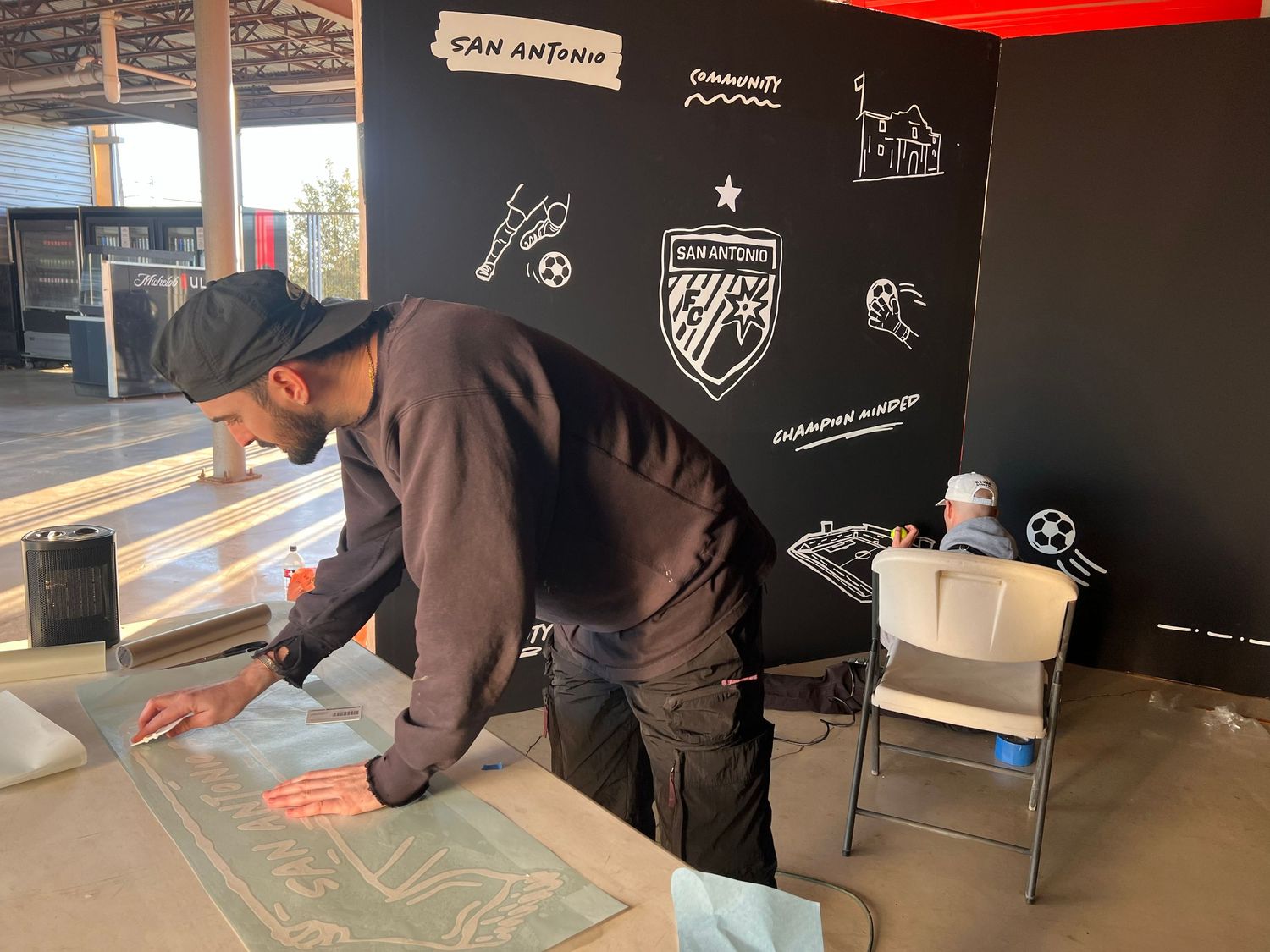 A person in dark clothing works on a project at a counter with a black wall featuring sports-themed graphics and logos.