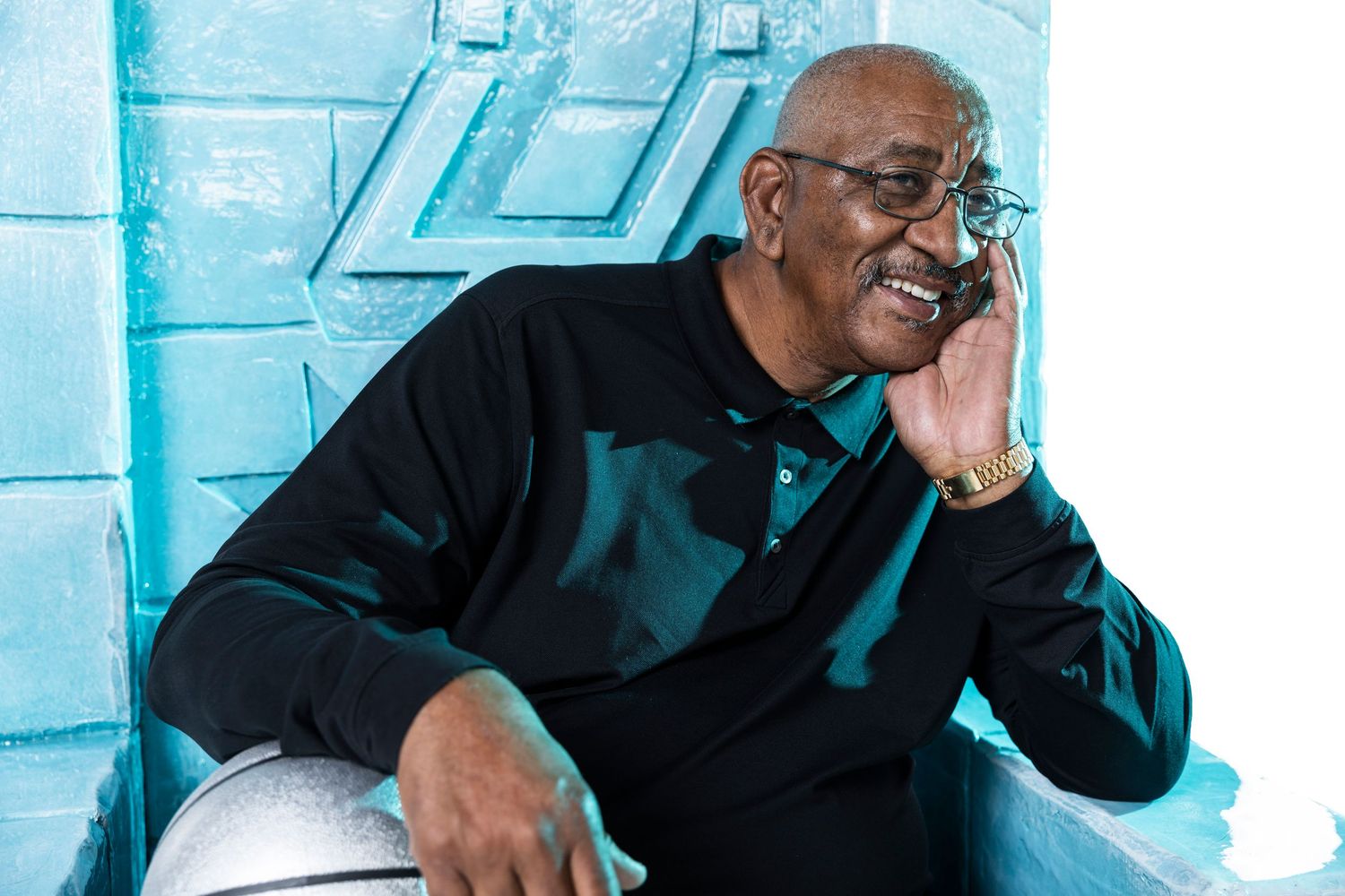 A person in a black outfit smiles and gestures while sitting against a blue-tinted background.