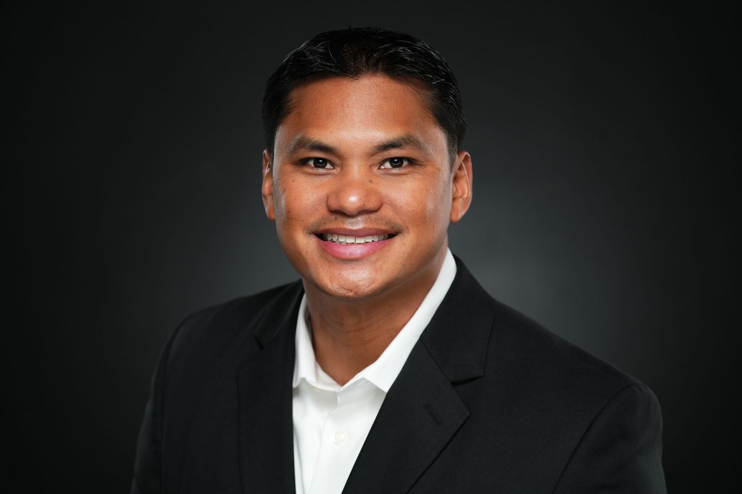 Professional headshot portrait of someone in a black suit and white dress shirt against a dark gray background.