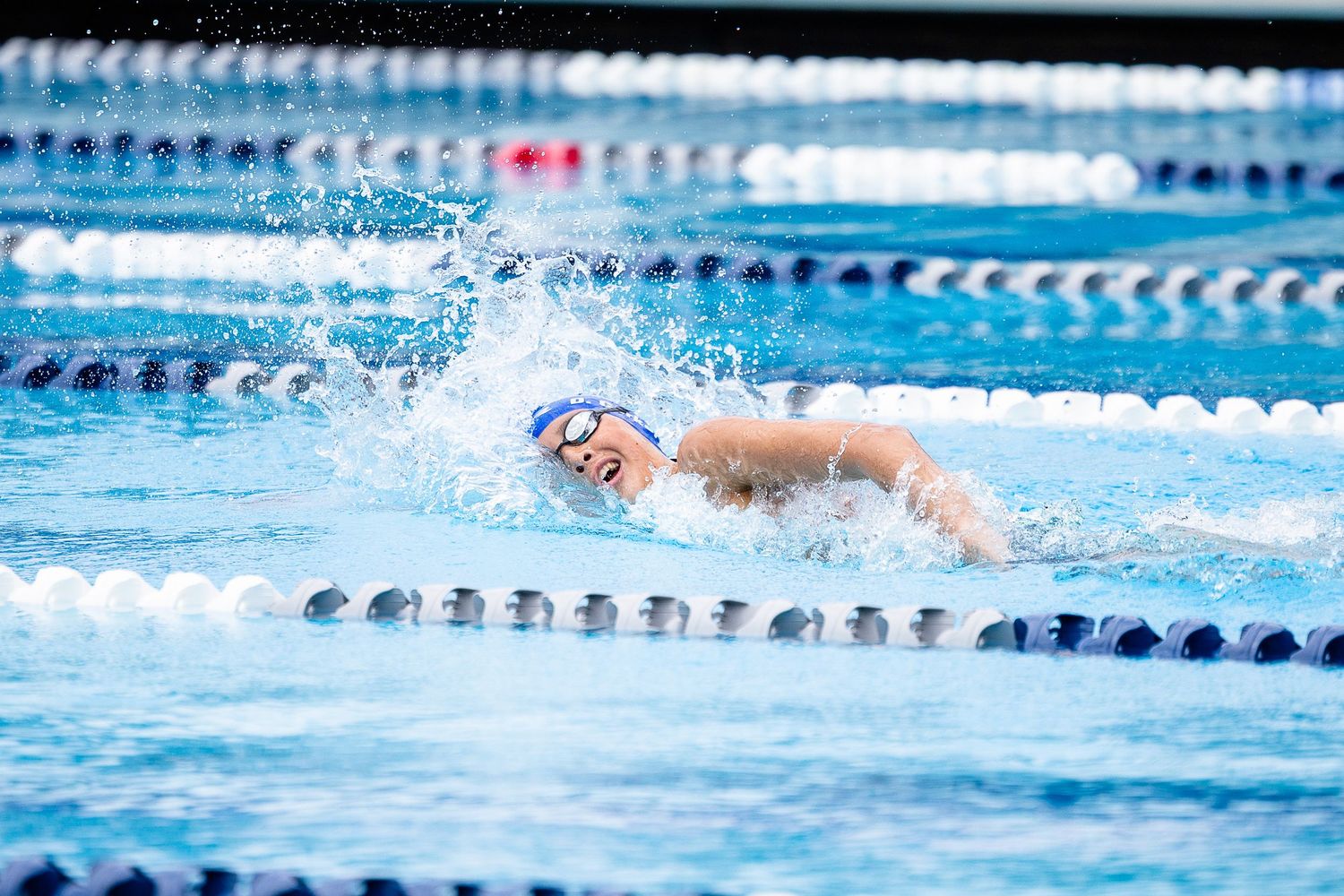 Competitive swimmer in a blue cap and goggles performs the freestyle stroke in a pool lane, splashing water beside floating lane dividers.