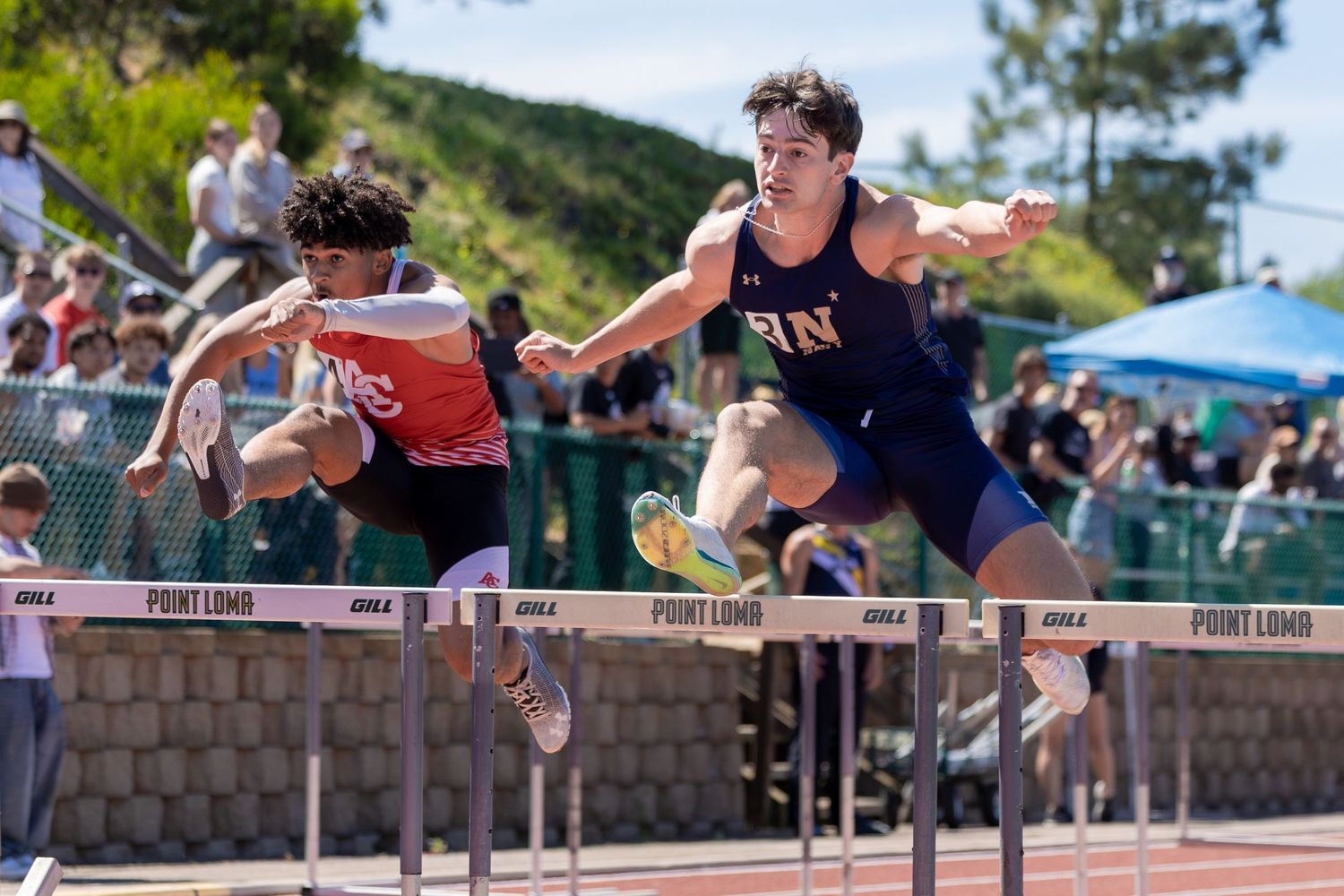 Two high school male athletes leap over hurdles during a track and field race while spectators watch from the stands on a sunny day.