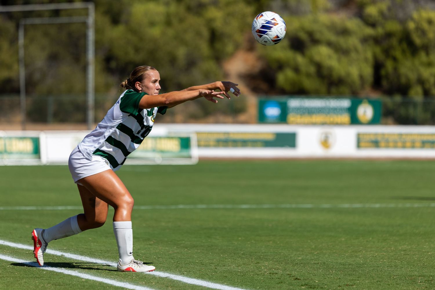 Female soccer player in a green and white striped uniform performs a throw-in on a grass field as the ball flies overhead.