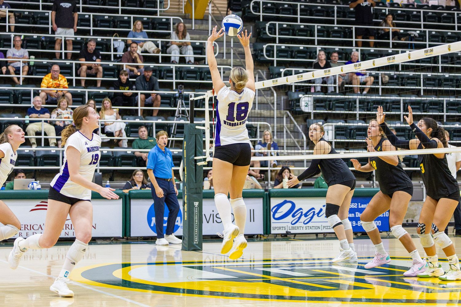 Volleyball player in a purple uniform leaps to spike the ball at the net during an indoor college match as teammates and opponents look on.