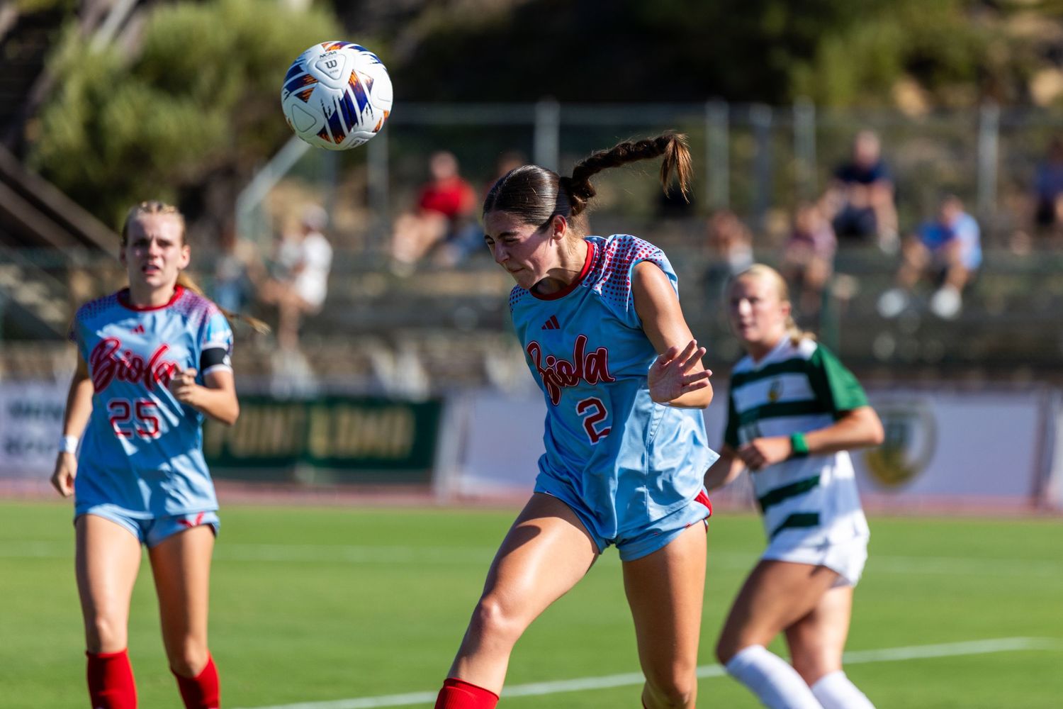 Female soccer player in a light blue uniform heads the ball during a college match while teammates and an opponent look on.