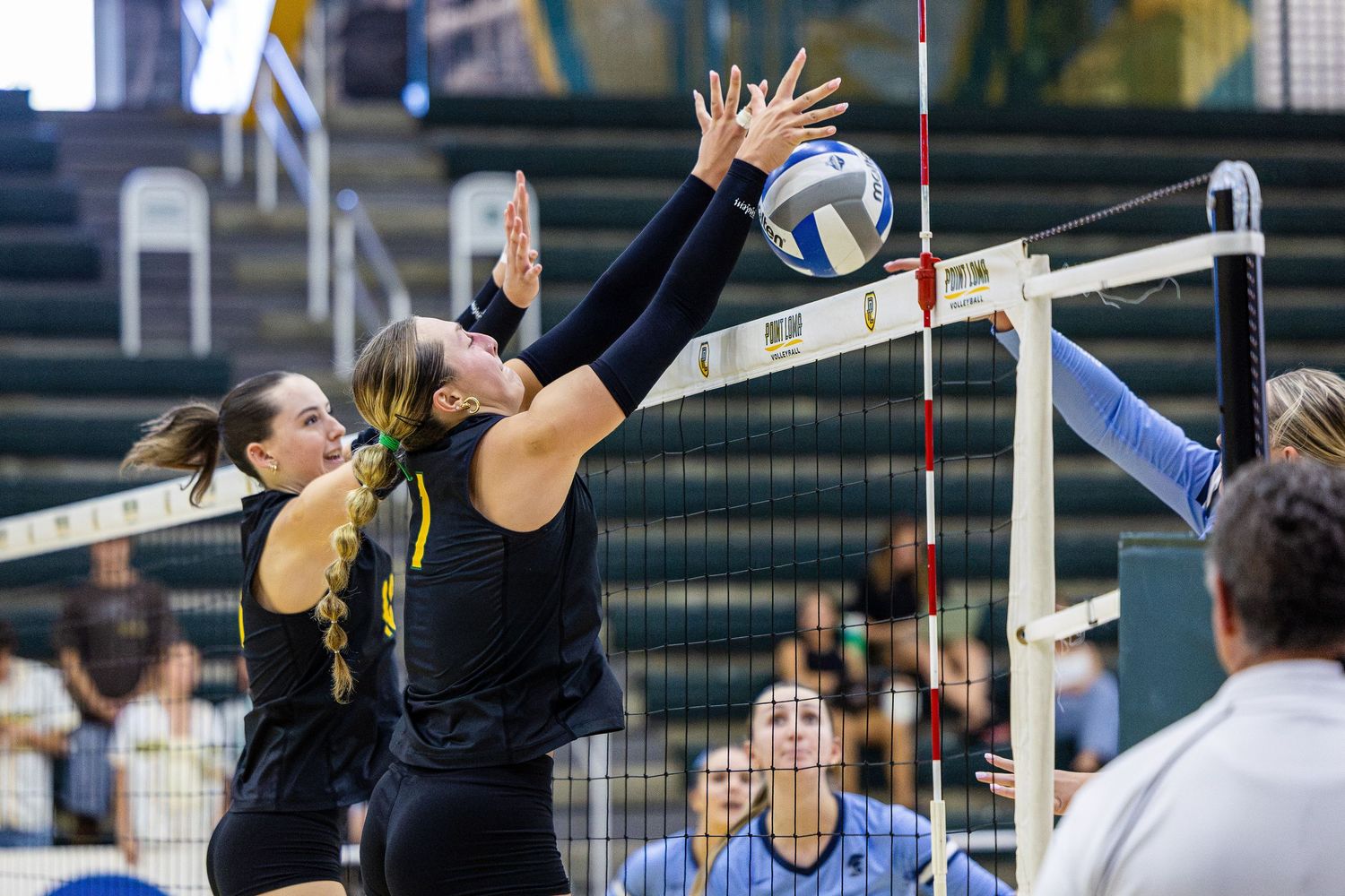 Two volleyball players in black jerseys jump at the net to block a spike during a college match as teammates and spectators watch.