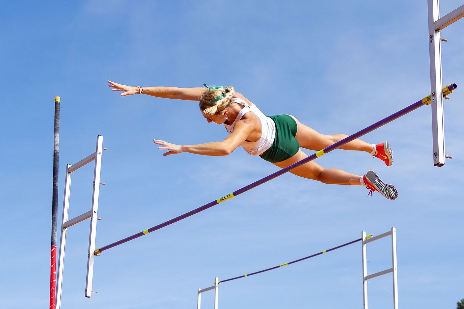 Female pole vaulter in a green and white uniform soars horizontally over the crossbar against a clear blue sky during a track meet.