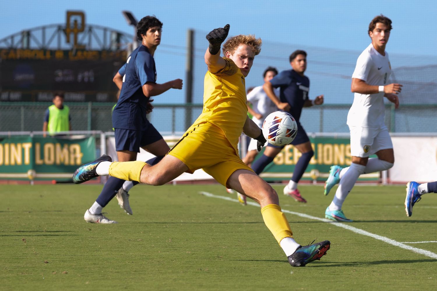 Soccer goalkeeper in a yellow kit stretches to kick the ball during a match while players in navy and white watch on a sunny field.
