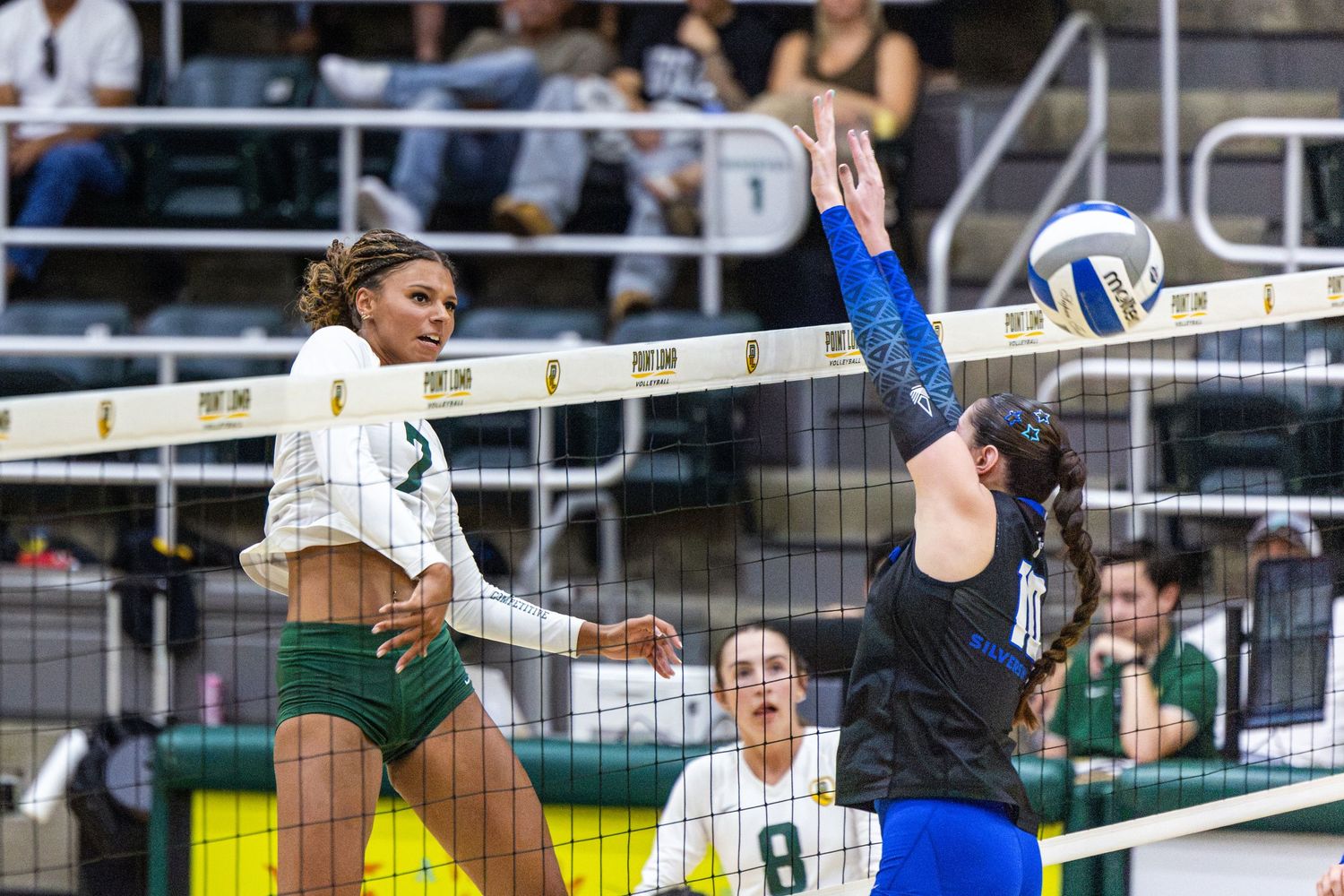 Volleyball players in green and blue uniforms leap at the net as a blocker reaches to deflect the ball during a college match.
