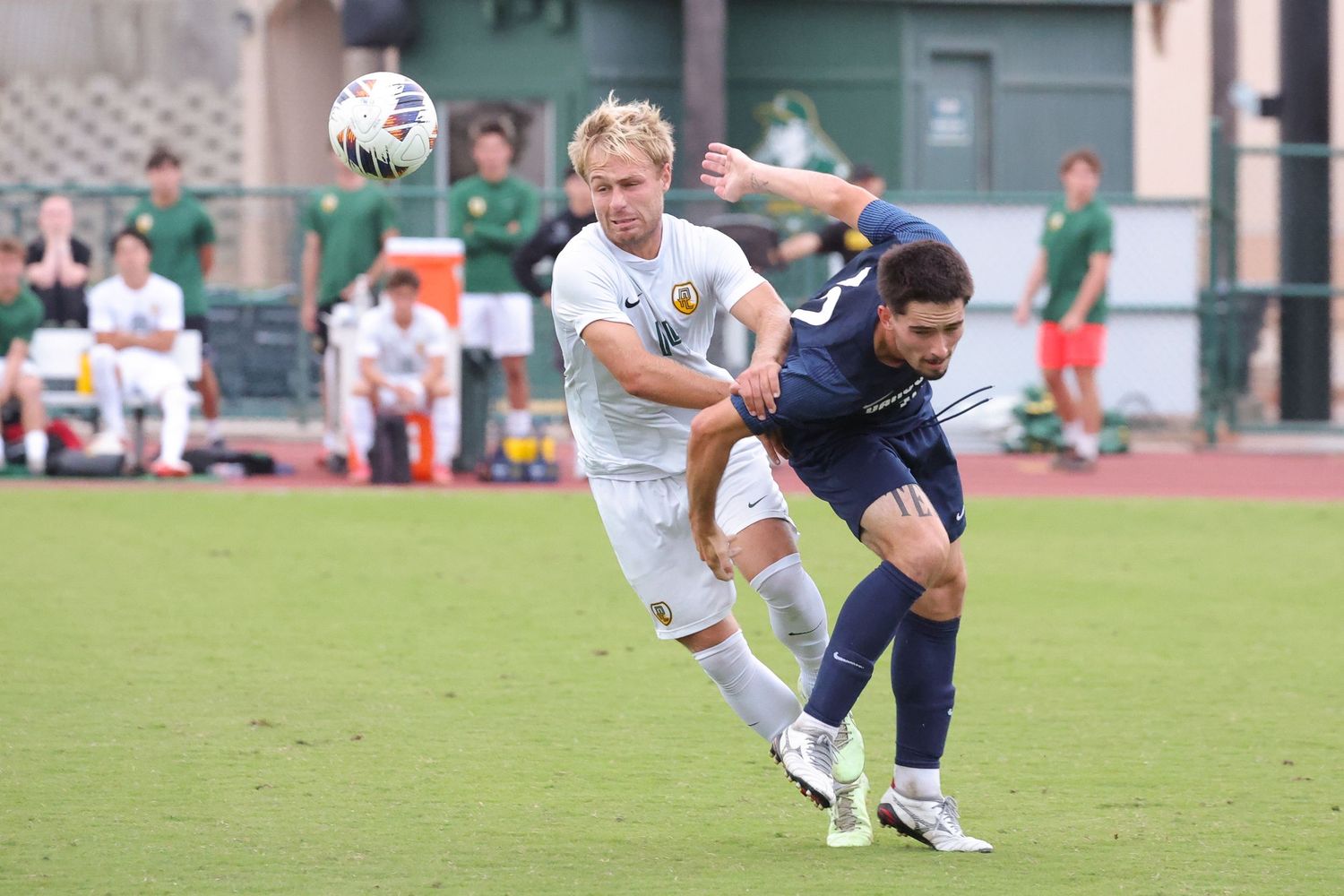 Two college soccer players battle for a header near the ball during a match while teammates watch from the sideline on a green field.