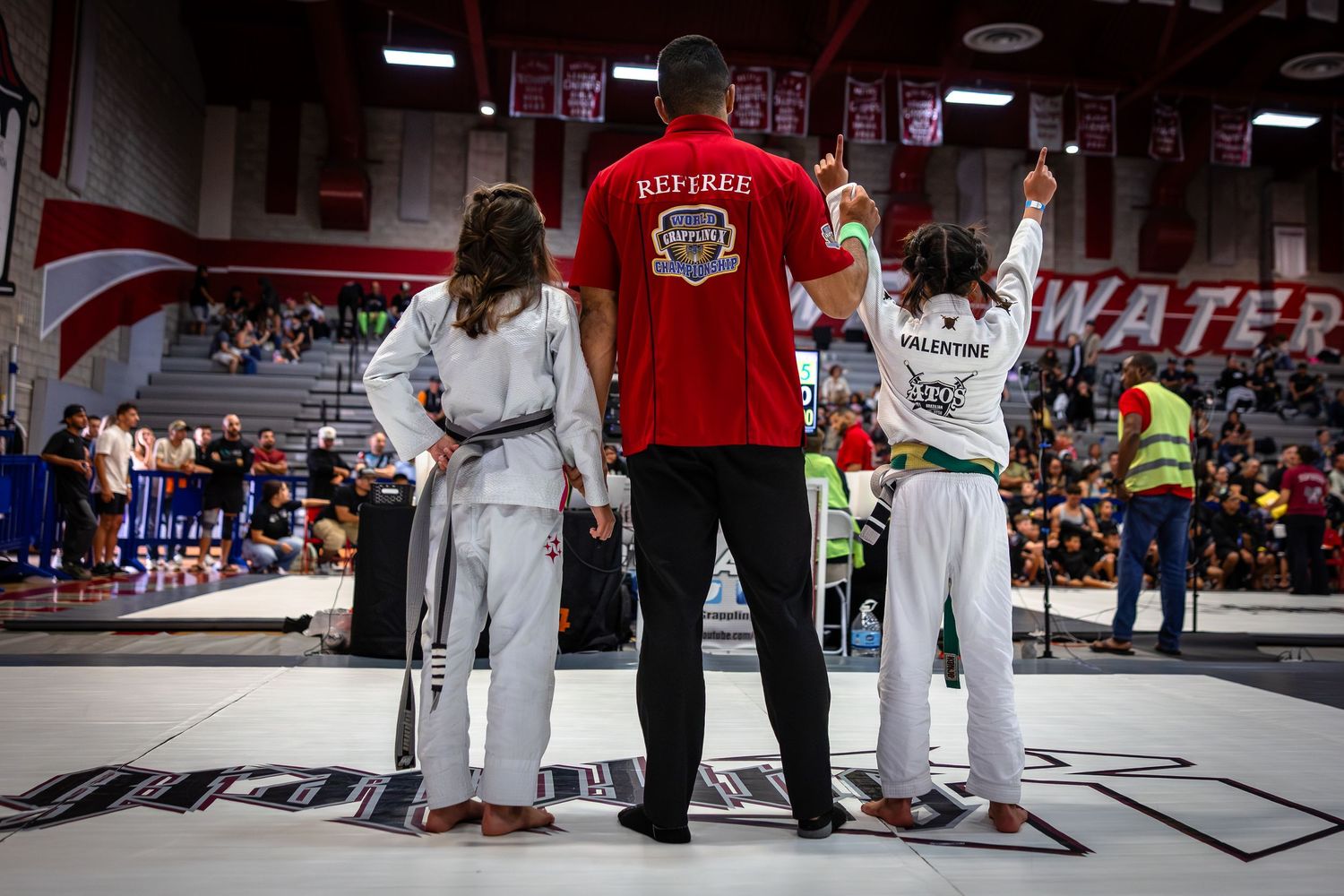 Two martial artists in white uniforms stand with referee on competition mat during tournament match.