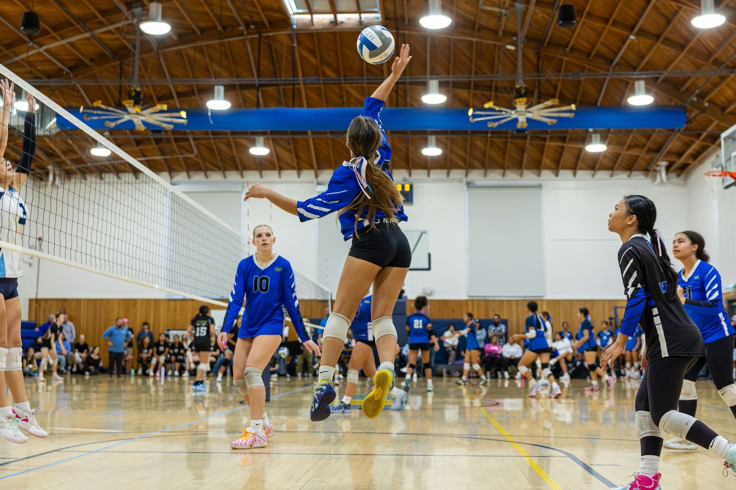 Volleyball player in a blue jersey leaps to spike the ball at the net during an indoor high school match as teammates and spectators watch.