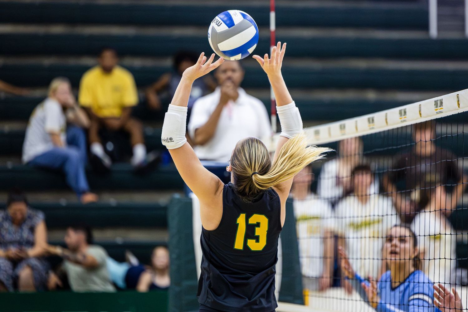 Female volleyball player in a black number 13 jersey jumps at the net to set the ball while spectators watch from the stands.