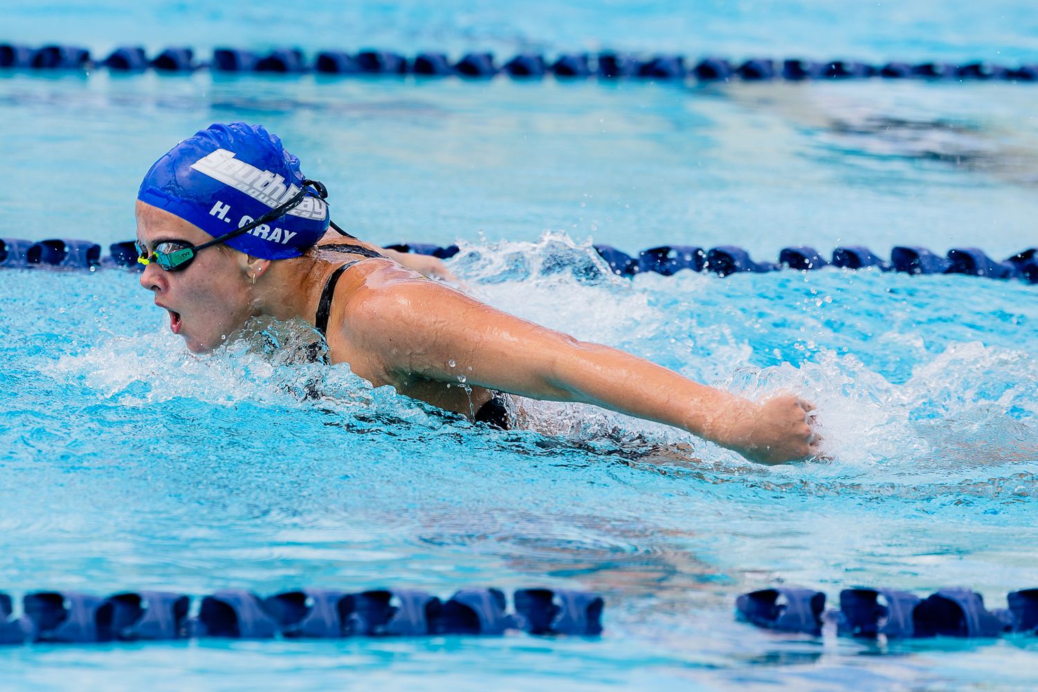 Female swimmer in a blue cap and goggles performs the butterfly stroke, arm extended forward through splashing water in a lane-lined pool.