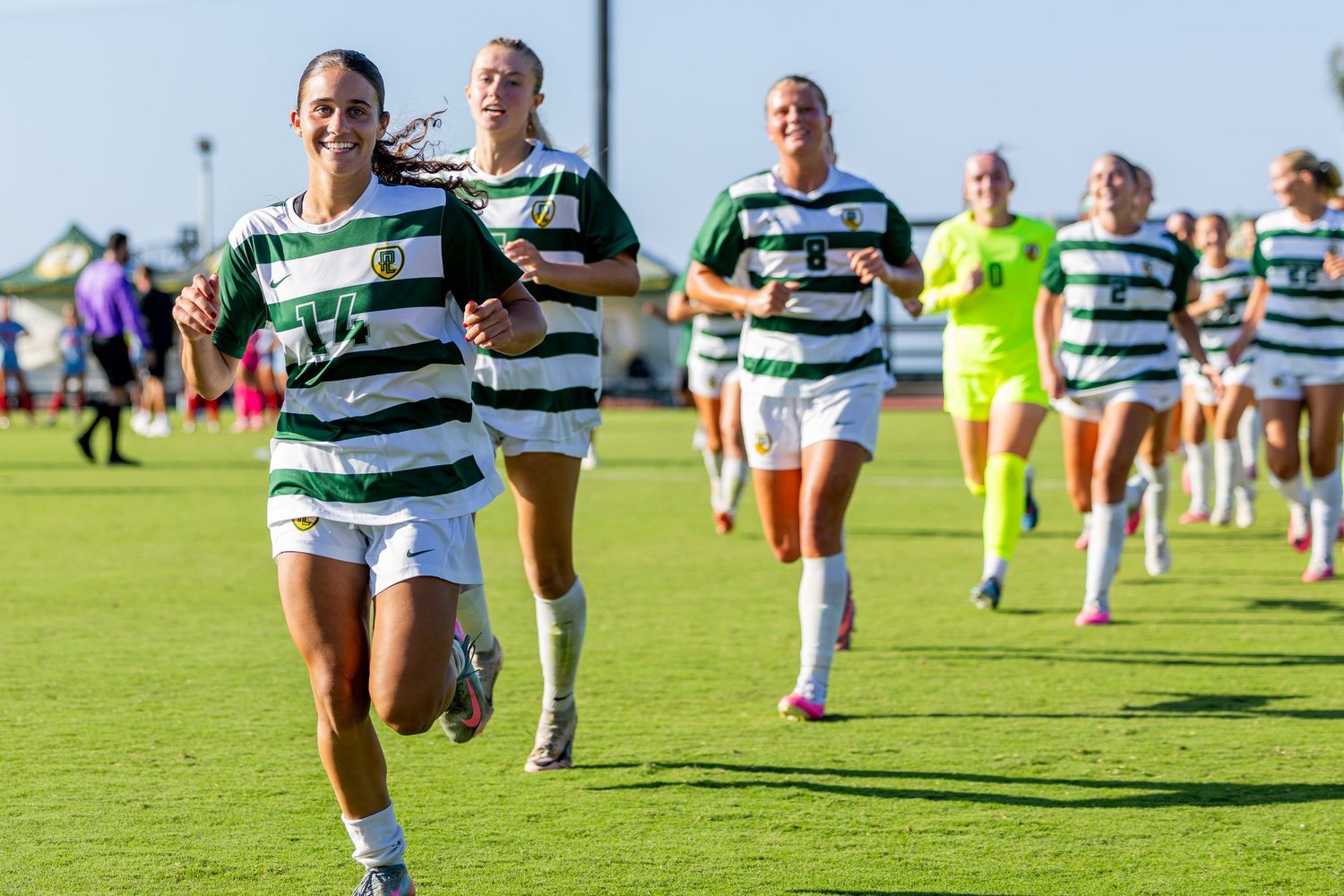 Women's soccer players in green and white striped jerseys jog across a sunny grass field while smiling during a college match.