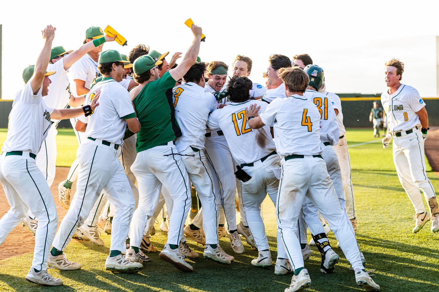 Baseball team celebrates victory in a group huddle on the field, wearing white and green uniforms.