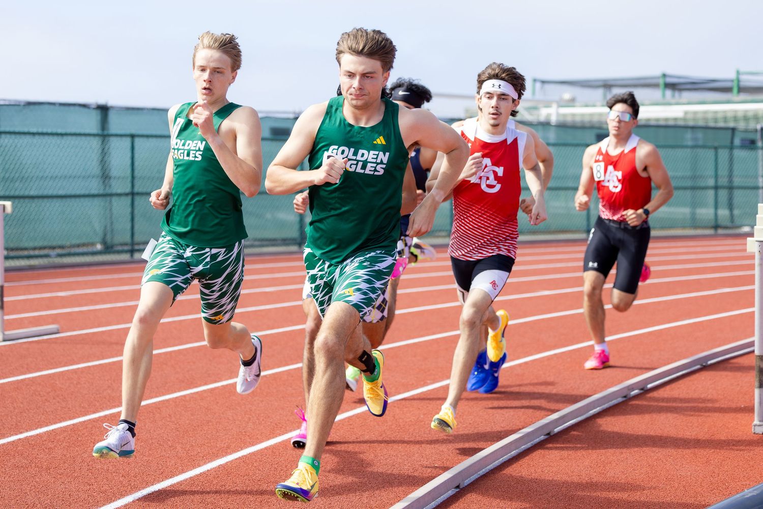 Four male high school runners in green and red uniforms race around a red outdoor track during a track and field meet.