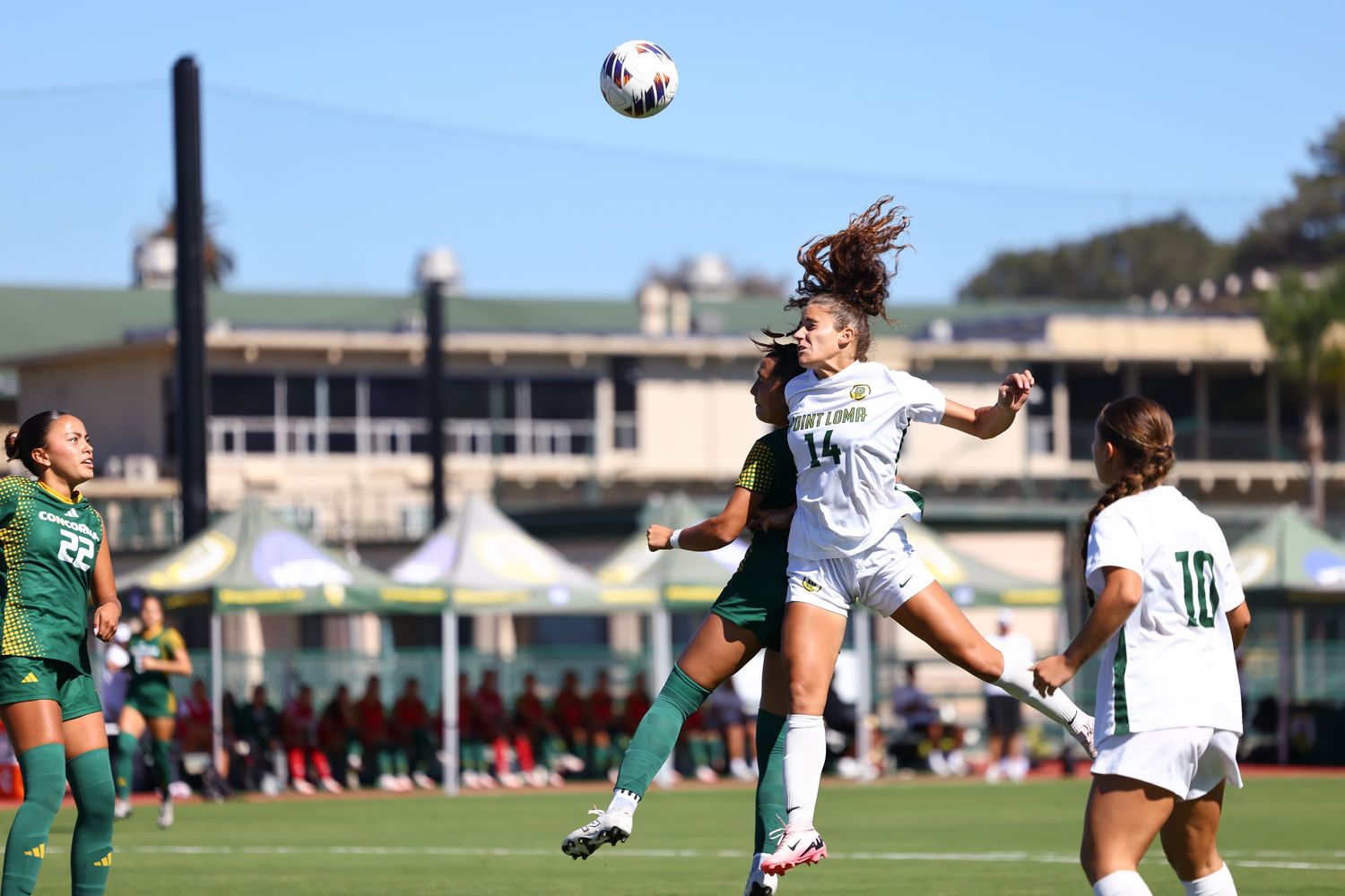 Two women's soccer players leap to head the ball during a match, with teammates and spectators watching from a grass field.