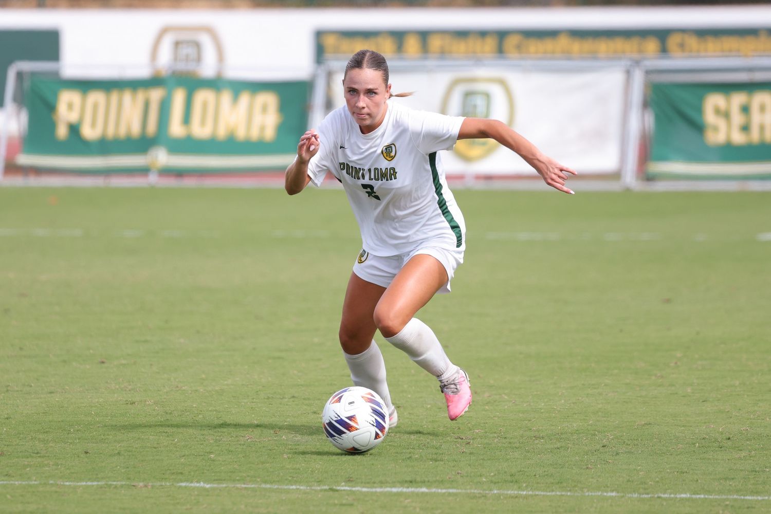 Female soccer player in a white Point Loma uniform dribbles the ball across a grass field during a college match.