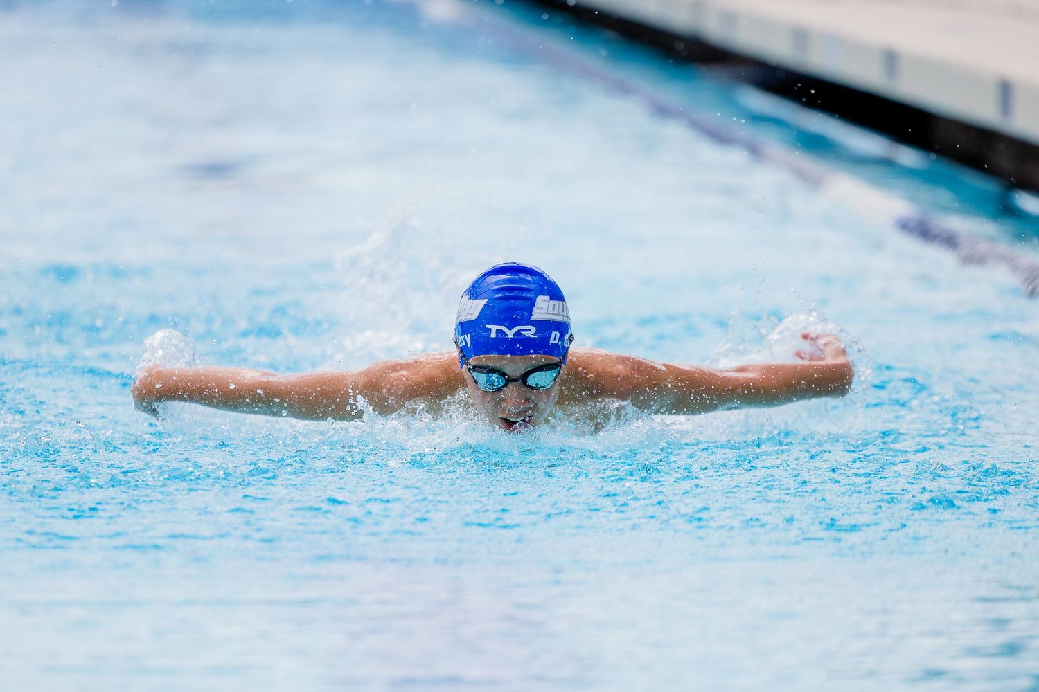 Swimmer in a blue cap and goggles performs the butterfly stroke with arms extended across the surface of a clear blue pool.