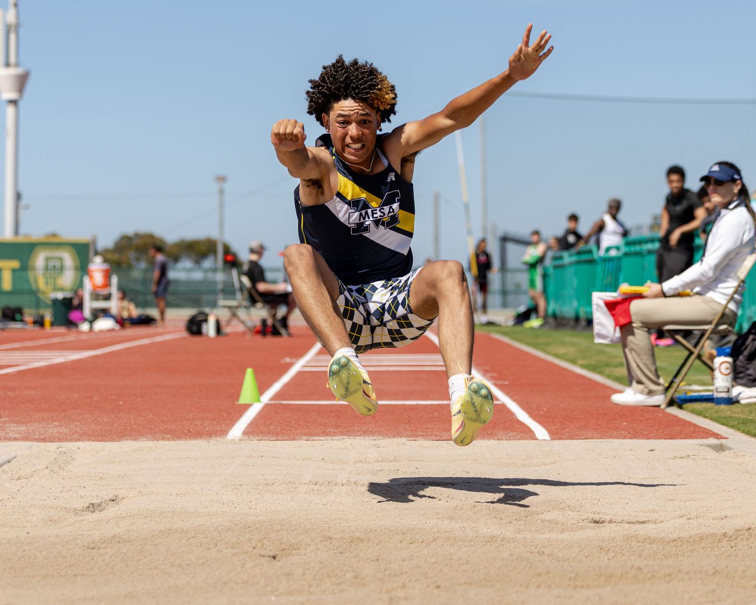 Long jumper in mid-air with arms outstretched above the sand pit during a track and field competition as spectators watch.