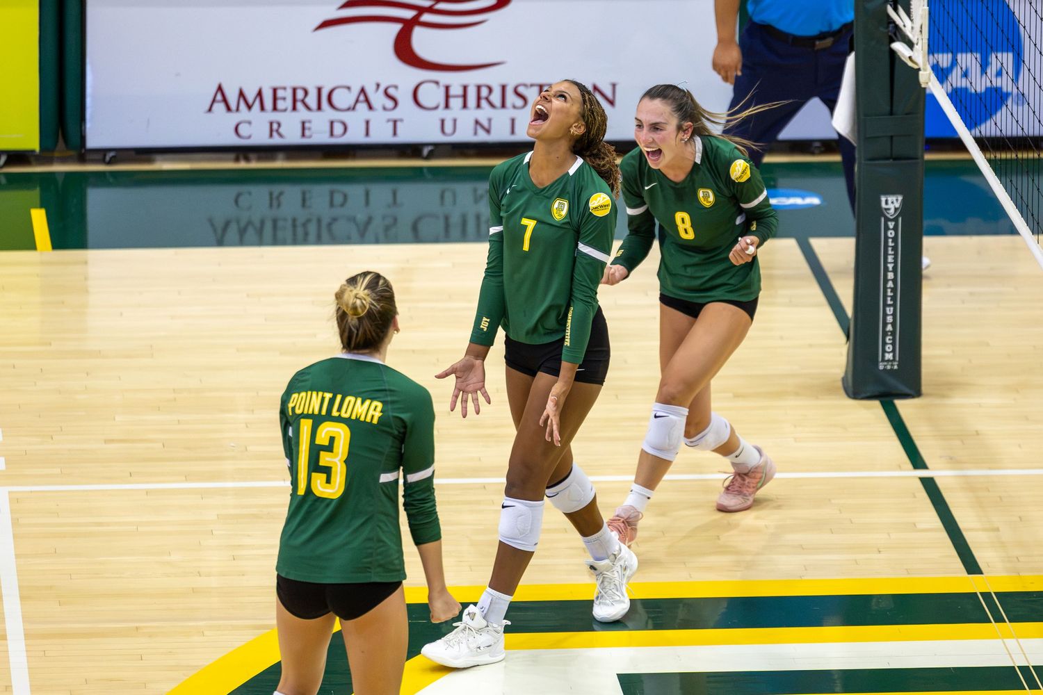 Three Point Loma women's volleyball players in green and gold uniforms celebrate and react excitedly during a match on an indoor court.