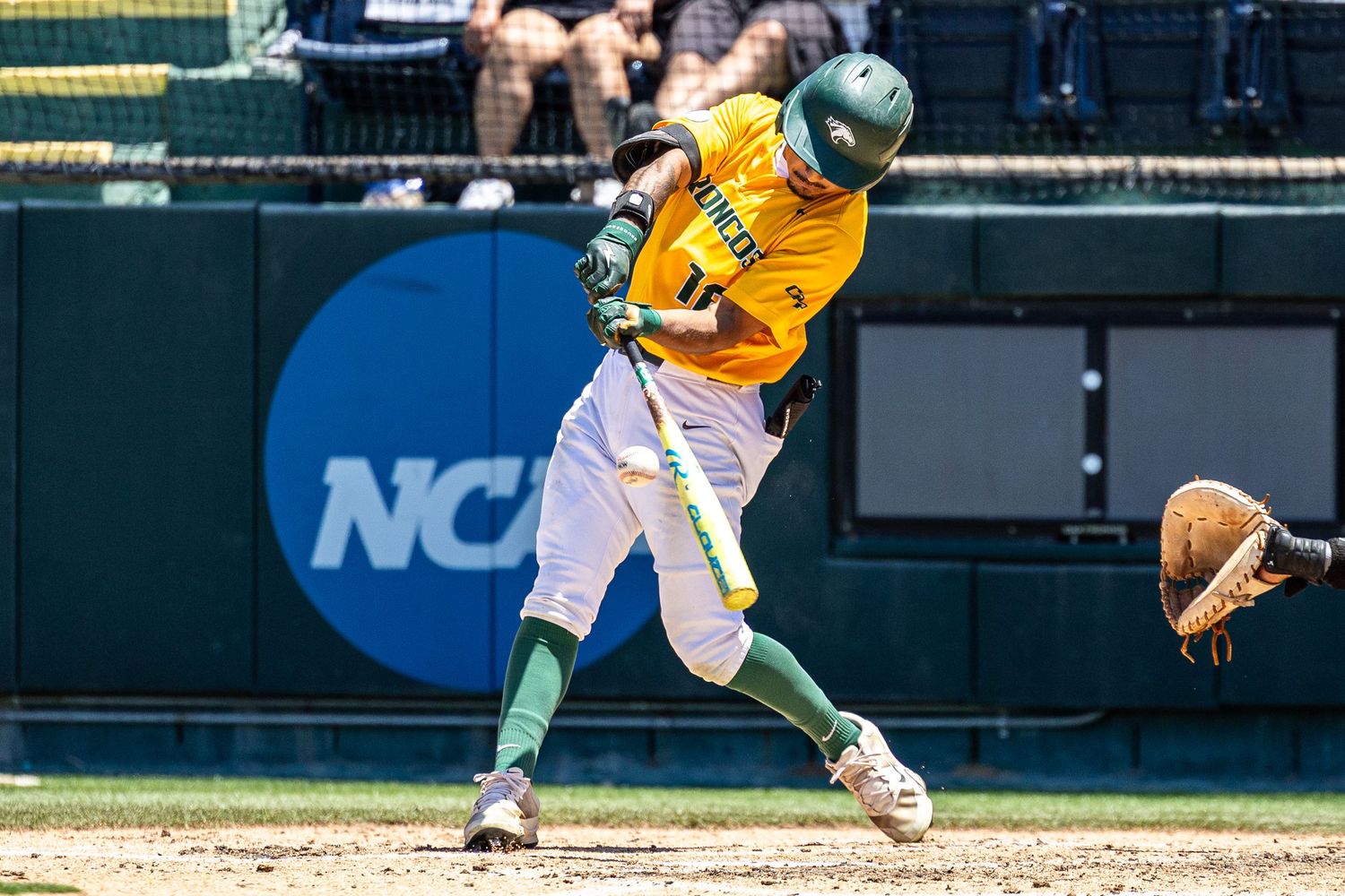 NCAA baseball player in yellow and green uniform swinging bat during a sunny collegiate game.