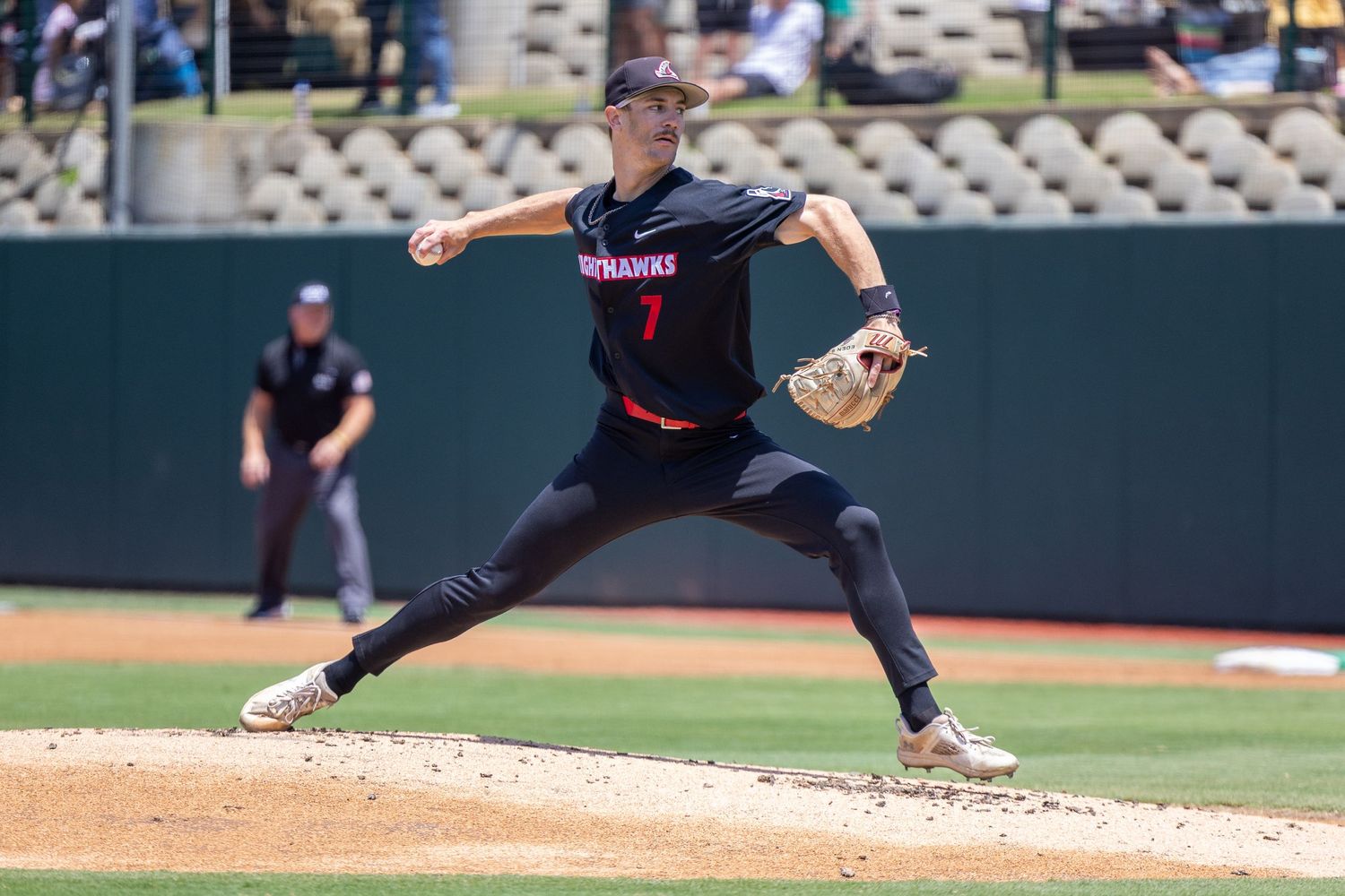 Baseball pitcher in a black number 7 jersey and gray pants mid-delivery on the mound while an umpire stands behind him on the field.