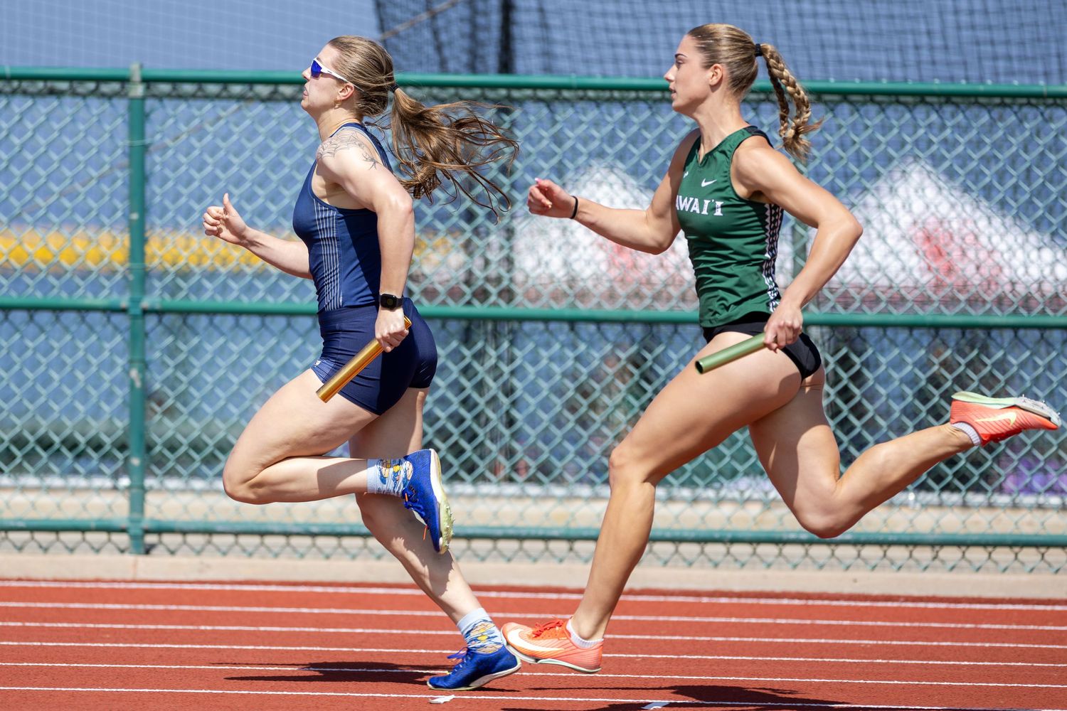 Two female sprinters in athletic uniforms race side by side down a red track during a track and field competition.