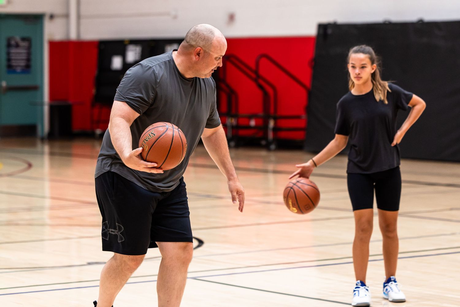 Two people practicing basketball drills together in a gymnasium with red walls.