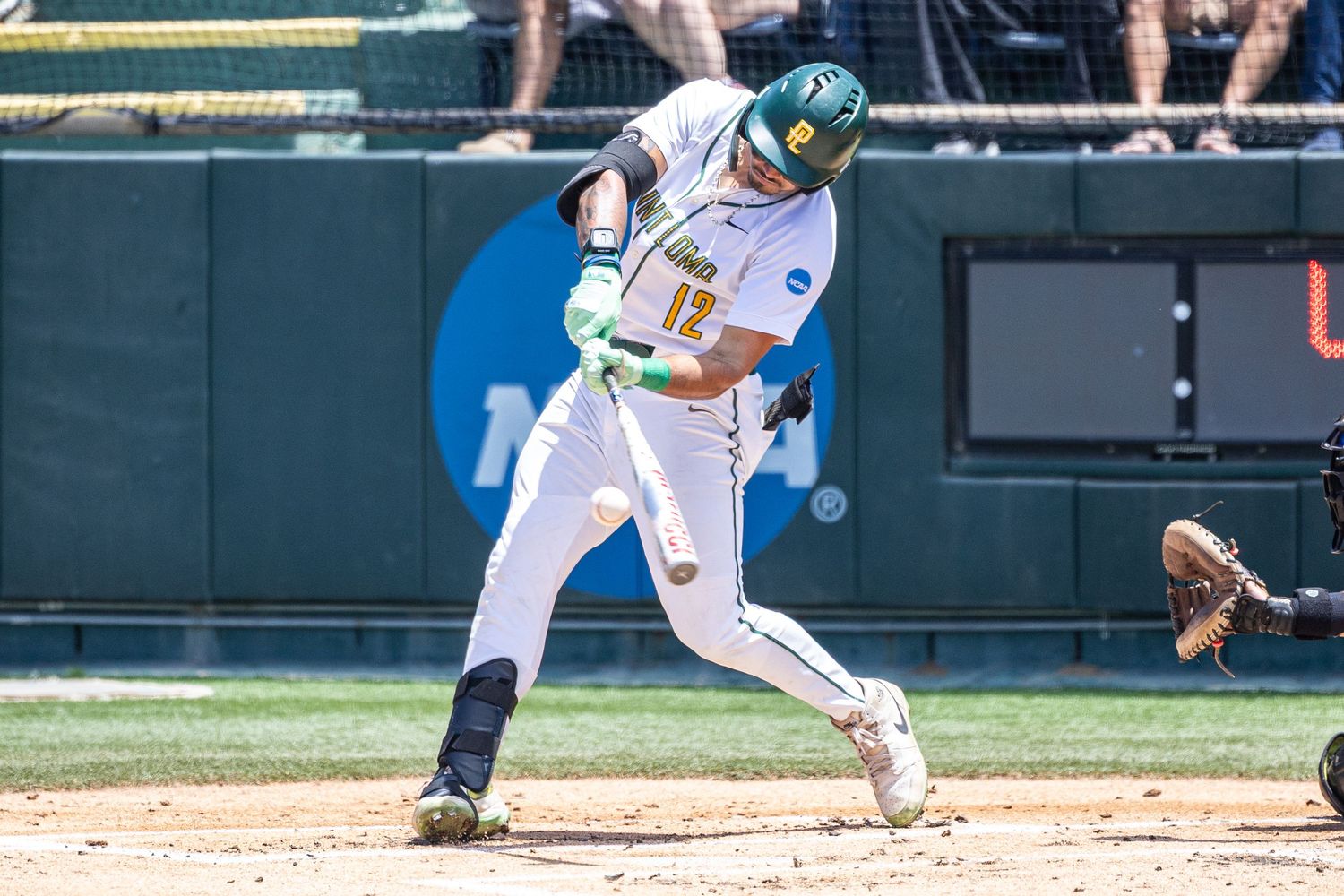 A college baseball batter in a green helmet and white uniform swings at a pitch during an NCAA tournament game as the catcher waits behind.