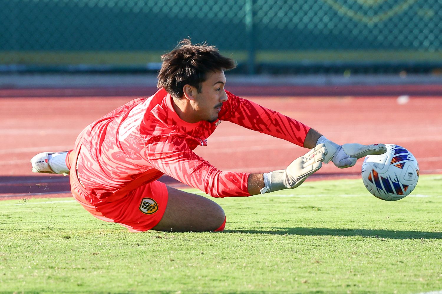 Goalkeeper in a red kit dives low on the grass to save a soccer ball during a match on the pitch.