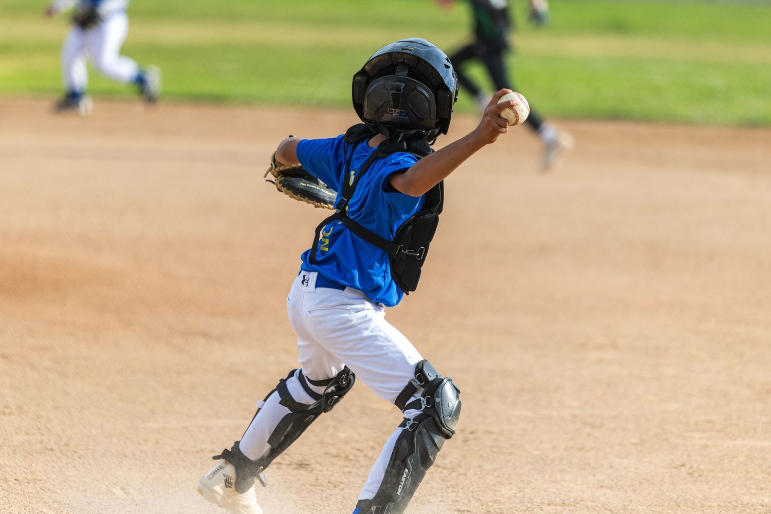 Young baseball catcher in a blue jersey, helmet and shin guards throws a ball on a dirt infield during a youth baseball game.