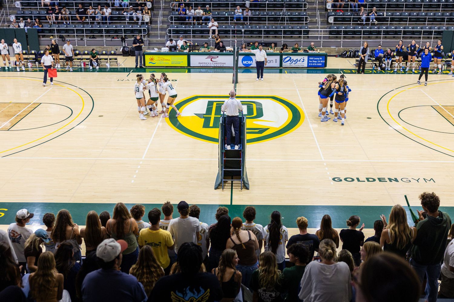 Volleyball teams stand on opposite sides of a gymnasium court with a green and gold center logo while fans watch from the foreground bleache