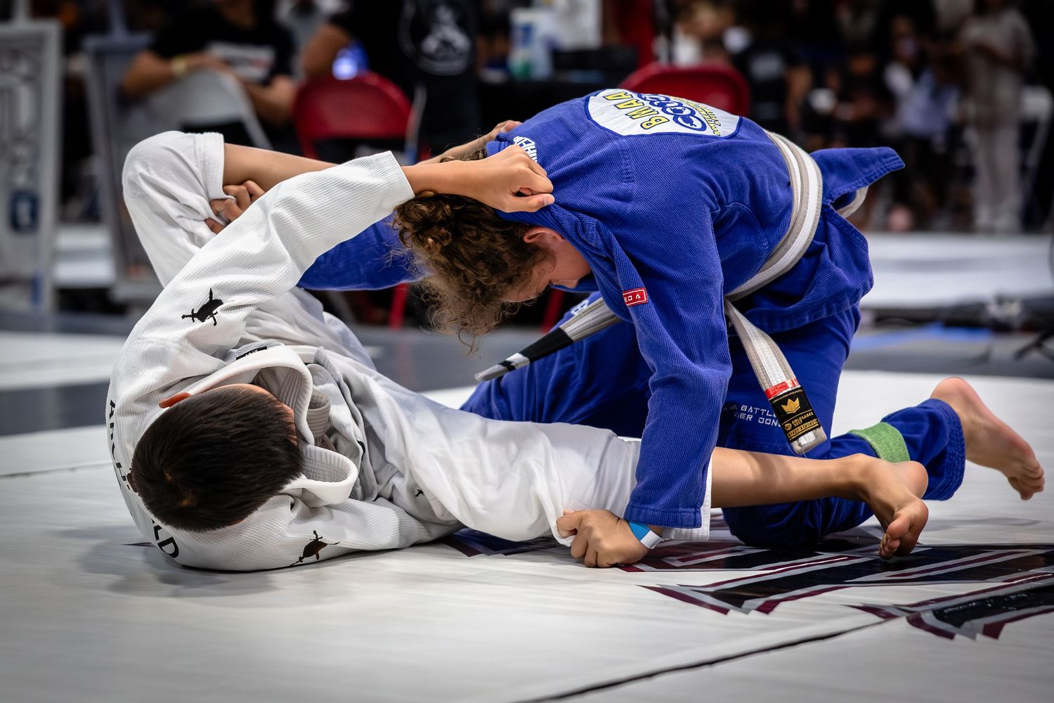 Two jiu-jitsu competitors grappling during a match on white mats.