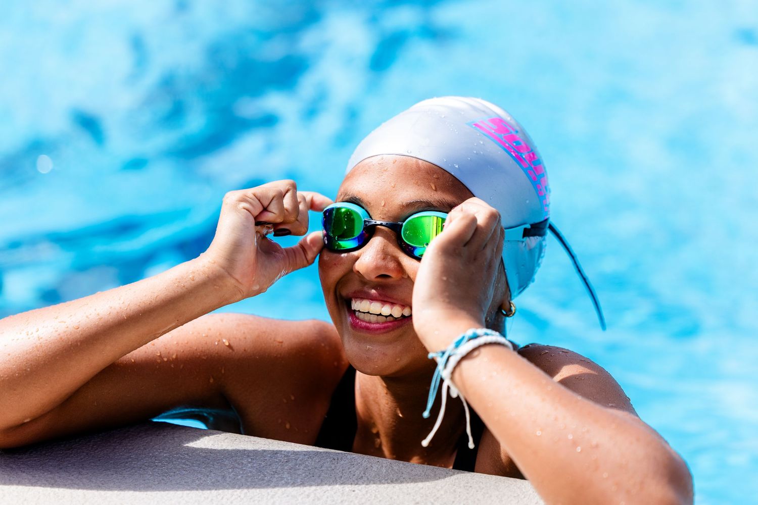 Smiling young girl in swim cap and green goggles resting at pool edge on a sunny day.