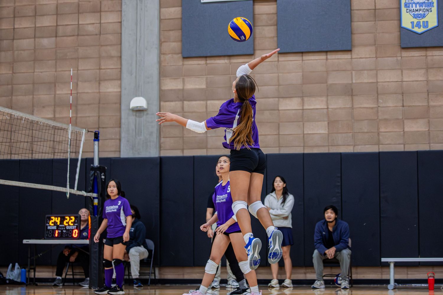 Indoor volleyball match with players in purple uniforms positioning for play.