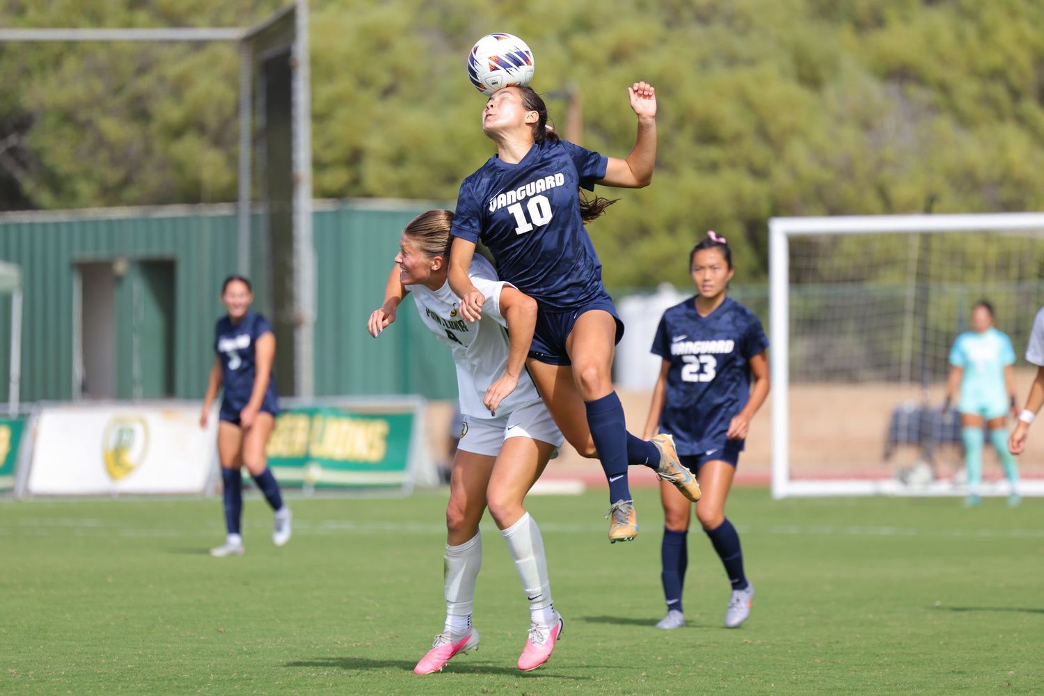 Two women's soccer players in navy and white uniforms leap to head the ball during a match while teammates look on from the grass field.