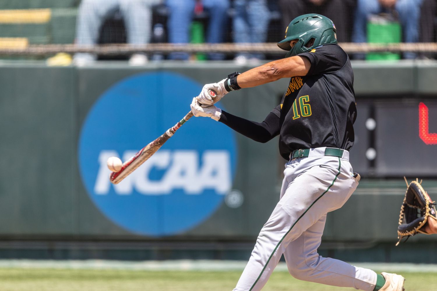 College baseball player in a green helmet and dark jersey swings at a pitch in front of an NCAA logo while the catcher reaches with a mitt.