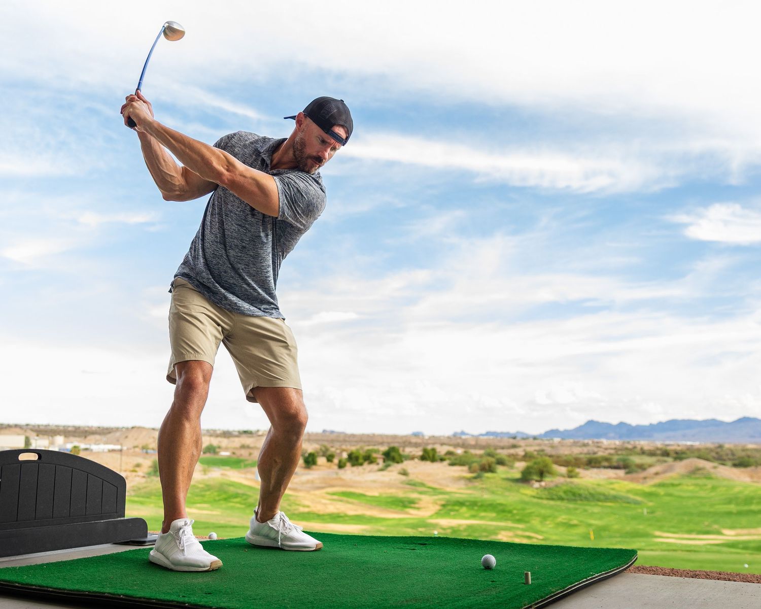Man swinging a golf club at a driving range with scenic desert mountains and blue sky in the background.