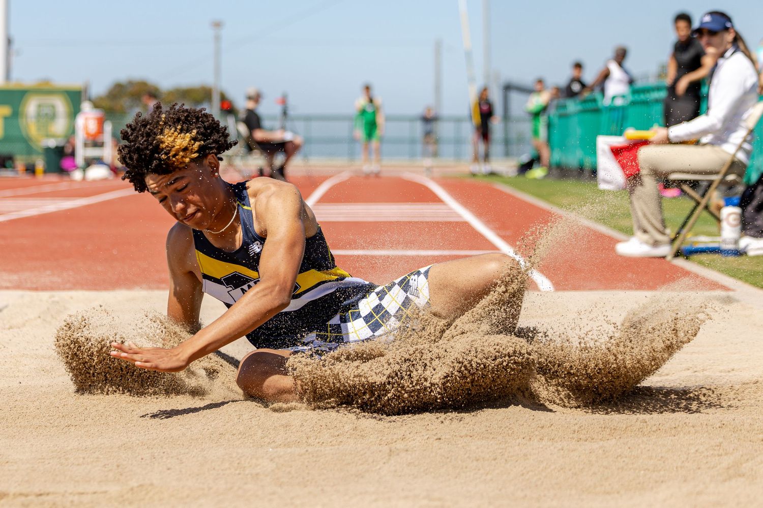 Track and field athlete lands in the sand pit during a long jump, kicking up sand on a sunny day as spectators watch from the sidelines.