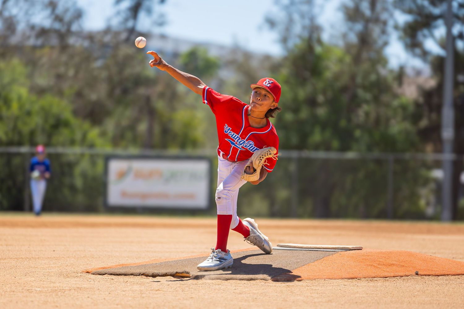 Youth baseball pitcher in red uniform throws pitch from mound during game.