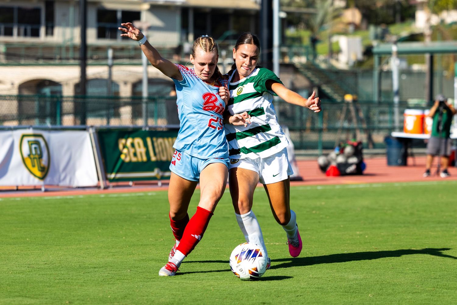 Two female soccer players in blue and green-striped uniforms battle for the ball during a college match on a sunny grass field.