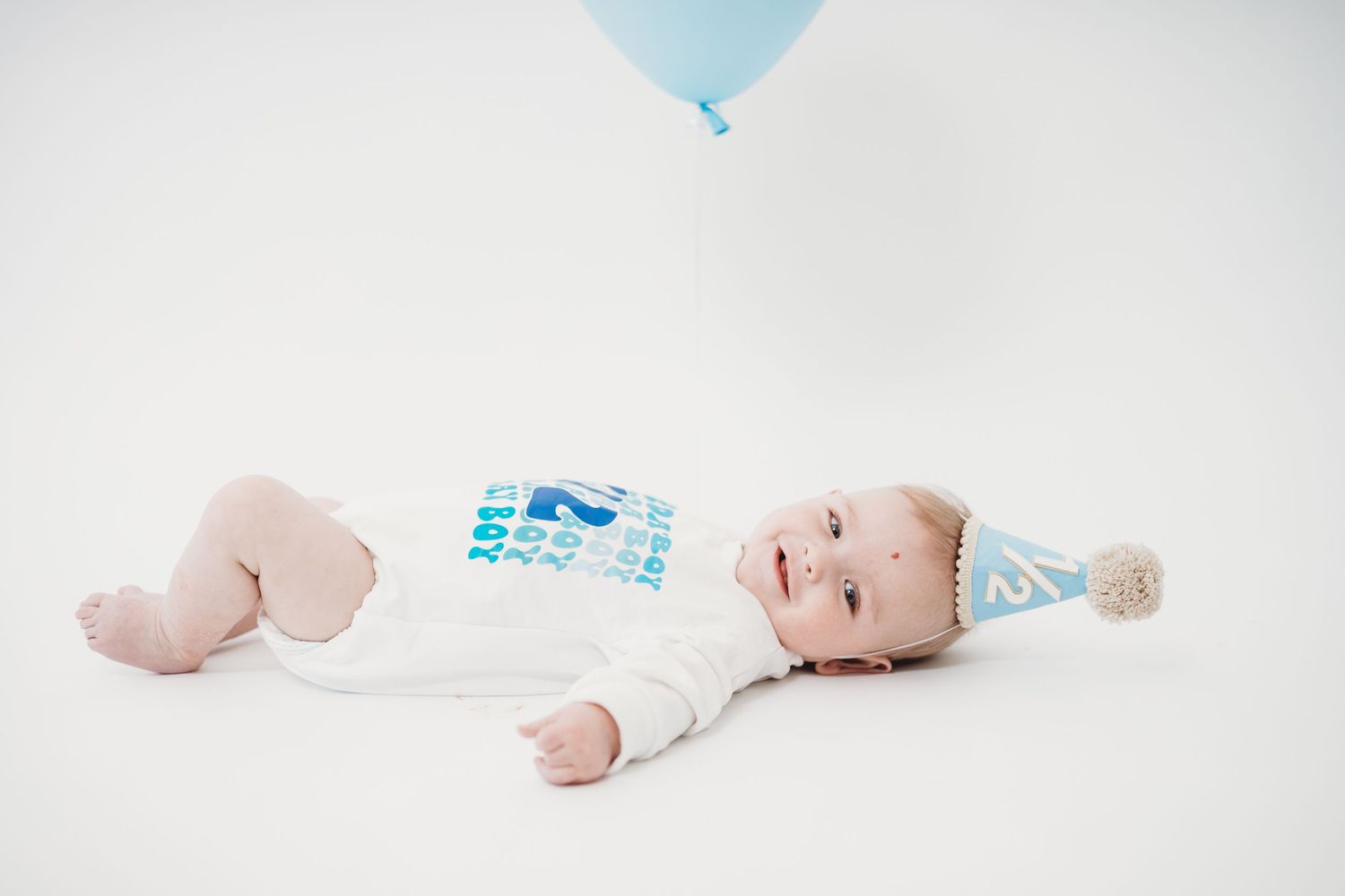 Baby lying on white backdrop with blue balloon and party decorations.