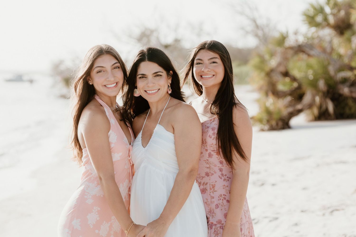 Three women in pink dresses laugh together on a sunny beach.