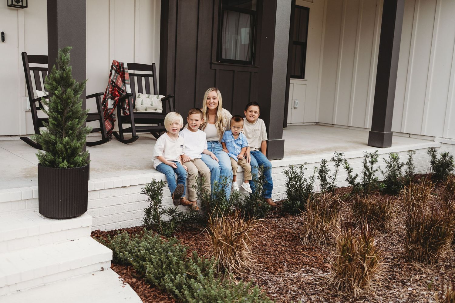 People sit on a porch of a white building with landscaping and mulch beds in the foreground.