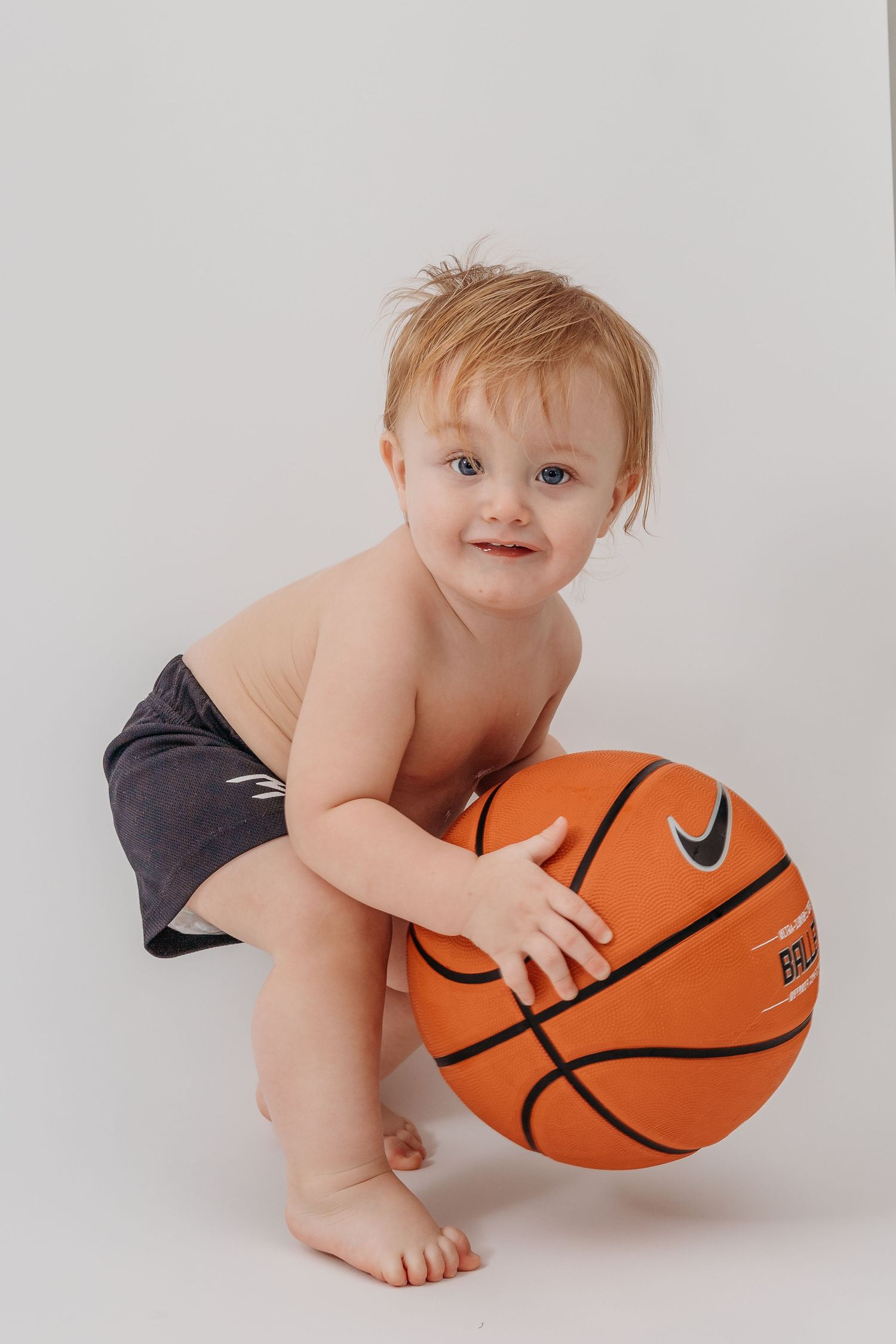 Baby in dark diaper playing with orange basketball on white background.