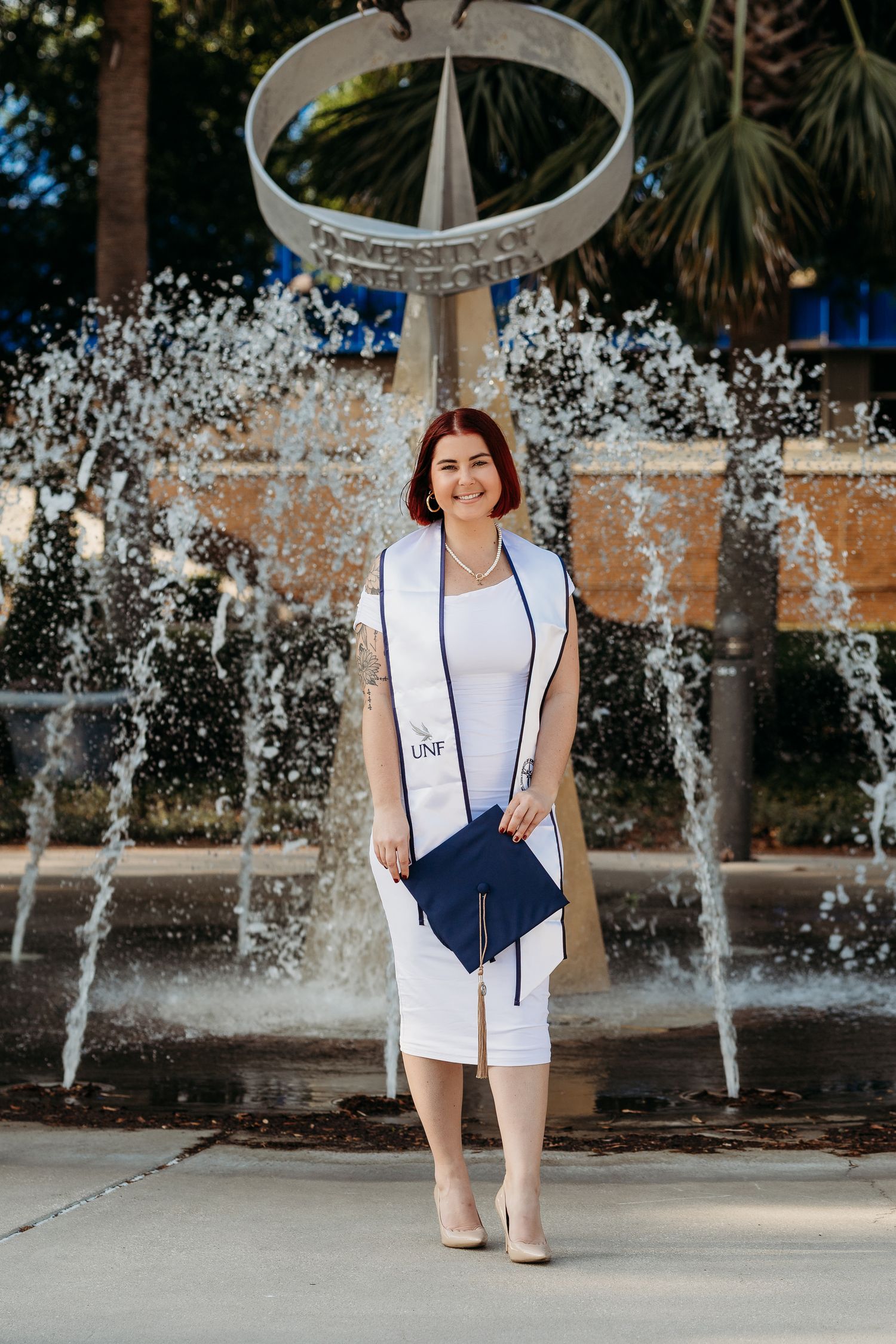Graduate in white dress standing in front of ornate fountain with water spraying.
