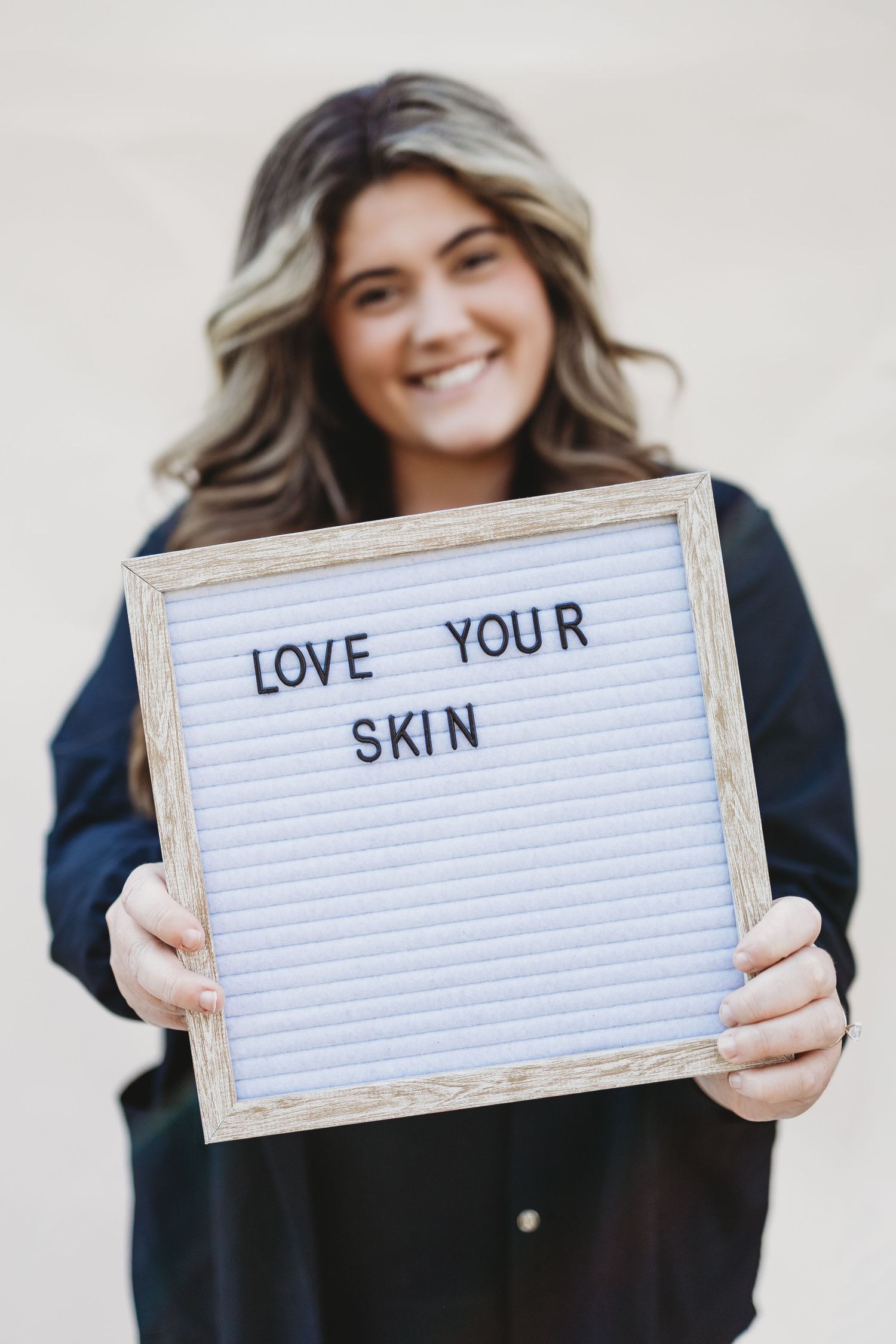 Someone holds a white letterboard sign with the text 'LOVE YOUR SKIN' while wearing a dark colored top.